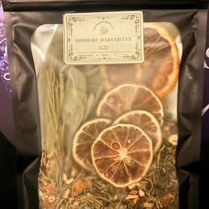 May include: A black resealable pouch containing a soy wax candle with dried citrus slices, herbs, and a label reading Midnight Margaritas. The clear window displays the botanical ingredients, including sliced limes and sage leaves inside the bag.