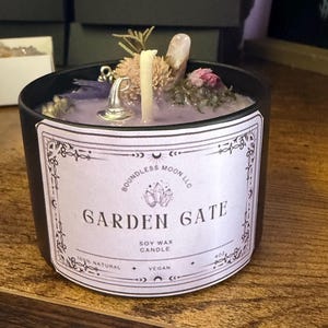 May include: A black tin soy wax candle with a light purple surface topped with dried flowers, a small silver witch hat charm, and a crystal. The label reads Boundless Moon LLC, Garden Gate, Soy Wax Candle, 100% Natural, Vegan, and 4oz.
