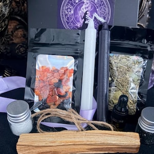May include: A ritual kit featuring a black box with a Boundless Moon label, one white candle, one black candle, two clear pouches containing orange stones and dried herbs, a wooden stick, a small dropper bottle, and two small jars with silver lids.