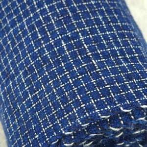 May include: A roll of deep blue fabric featuring a white grid pattern. The textile has a visible woven texture with small, light-colored horizontal and vertical dashes forming a consistent check design across the entire surface of the material.