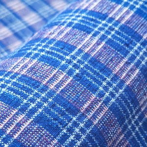 May include: A woven fabric featuring a vibrant blue plaid pattern with intersecting white and thin red lines. The textile has a visible textured weave with a consistent grid design throughout, showcasing a dense and durable construction.
