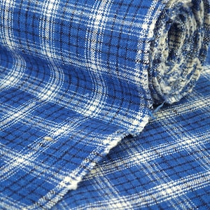 Puede incluir: Un rollo de tela tejida con un patrón de cuadros en azul, blanco y azul marino oscuro. El textil muestra un tejido de rejilla visible con líneas horizontales y verticales que se cruzan, creando una apariencia texturizada en toda la superficie.