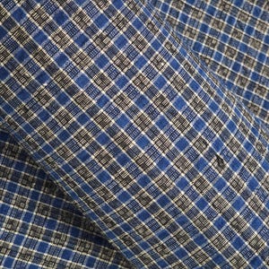 Op de afbeelding: Close-up van een stof met een blauw, zwart en wit ruitpatroon. De stof is gevouwen en toont de textuur en het ontwerp. De ruit bestaat uit vierkanten en rechthoeken.
