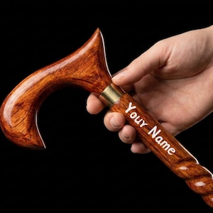 Könnte beinhalten: Ein polierter Gehstock aus Holz mit einem geschwungenen Griff und einem gedrehten Schaft. Der Stock verfügt über eine goldfarbene Metallmanschette, die den Griff mit dem Schaft verbindet, und den weißen Schriftzug Your Name auf dem Holz.