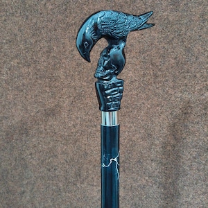 May include: A black walking cane featuring a handle shaped like a raven perched atop a human skull, which is held by a skeletal hand. The shaft of the cane is black with white lightning-like streaks and a silver-colored metal collar below the handle.