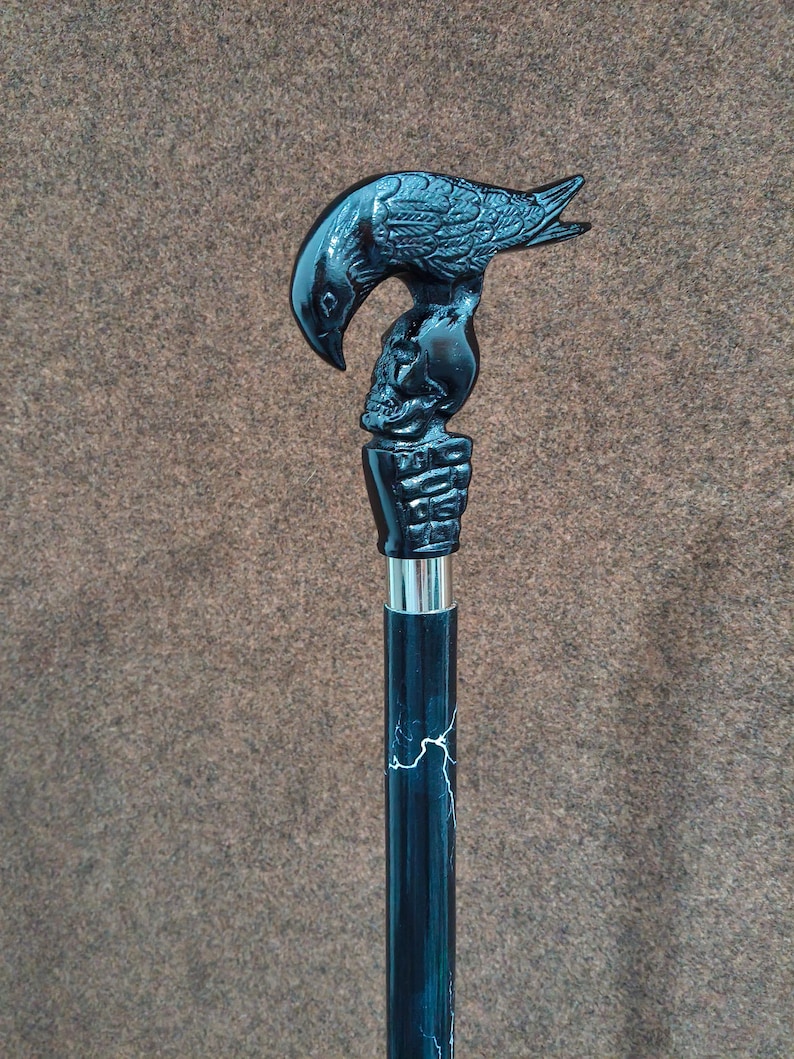 May include: A black walking cane featuring a handle shaped like a raven perched atop a human skull, which is held by a skeletal hand. The shaft of the cane is black with white lightning-like streaks and a silver-colored metal collar below the handle.