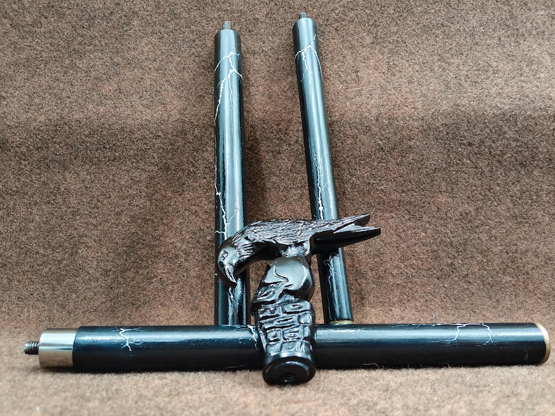 May include: A black walking stick handle featuring a carved raven perched atop a skull. The handle is attached to a black shaft with white marble-like veining. Two additional matching shaft segments are positioned vertically behind the handle.