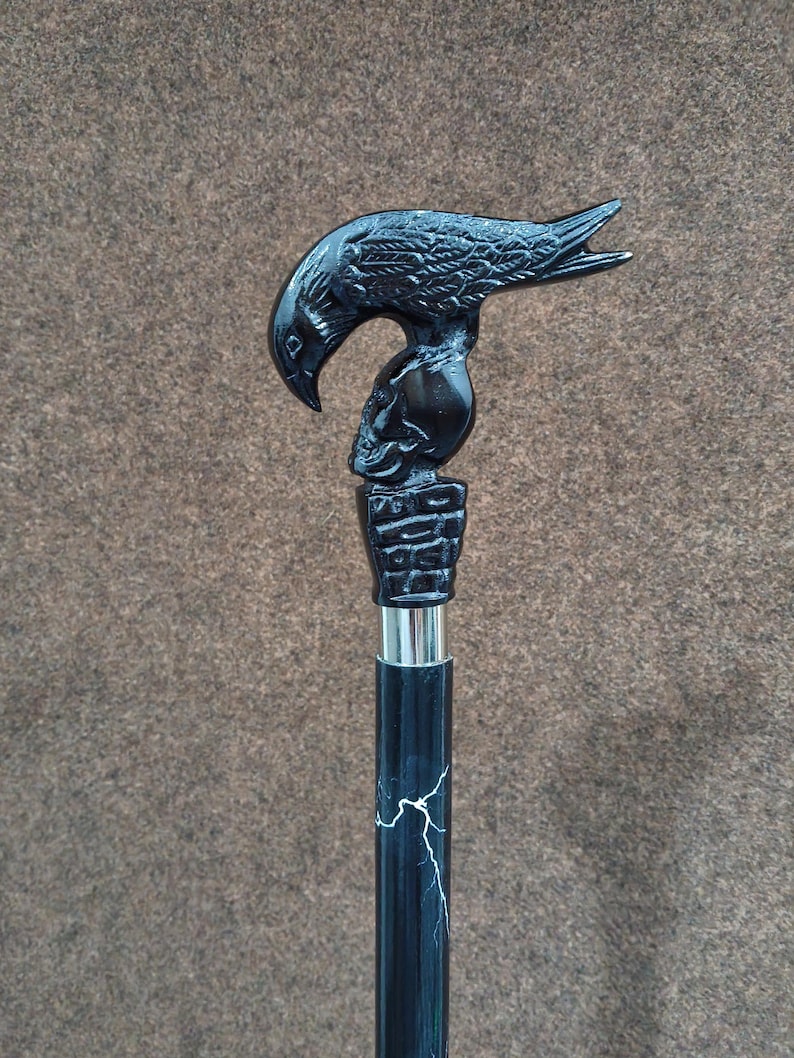 May include: A black walking cane featuring a handle shaped like a raven perched atop a human skull, which rests on a brick-patterned base. The shaft is black with a white lightning bolt pattern and includes a silver-colored metal collar connector.