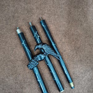 May include: Three black cylindrical segments of a walking stick featuring a white lightning bolt pattern. A black raven figure is attached to the central segment, with a metallic threaded connector at the top of each piece and a metal ferrule at the base.