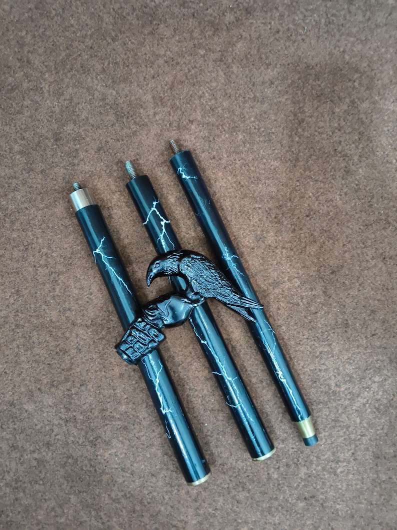 May include: Three black cylindrical segments of a walking stick featuring a white lightning bolt pattern. A black raven figure is attached to the central segment, with a metallic threaded connector at the top of each piece and a metal ferrule at the base.