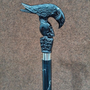 May include: A walking cane featuring a black handle shaped like a raven perched atop a human skull, which rests on a rectangular base. The shaft is black with a white lightning bolt pattern and is connected to the handle by a silver metal collar.