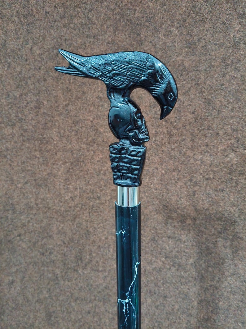 May include: A walking cane featuring a black handle shaped like a raven perched atop a human skull, which rests on a rectangular base. The shaft is black with a white lightning bolt pattern and is connected to the handle by a silver metal collar.