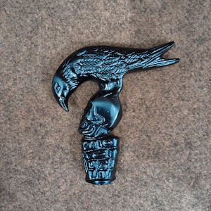 May include: A black cane handle featuring a sculpted raven perched atop a human skull, which rests on a textured, cylindrical base. The entire piece is finished in a solid, glossy black color with detailed carvings of feathers and bone.