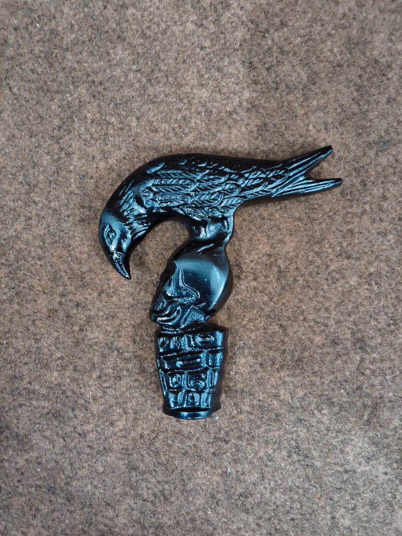 May include: A black cane handle featuring a sculpted raven perched atop a human skull, which rests on a textured, cylindrical base. The entire piece is finished in a solid, glossy black color with detailed carvings of feathers and bone.