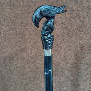 May include: A black walking cane featuring a detailed handle shaped like a raven perched atop a human skull, which is held by a skeletal hand. The shaft is black with white lightning-like streaks and a silver-colored metal collar below the handle.
