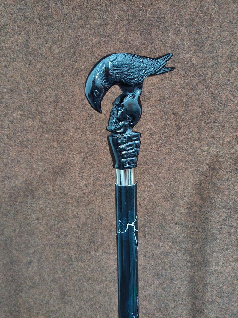 May include: A black walking cane featuring a detailed handle shaped like a raven perched atop a human skull, which is held by a skeletal hand. The shaft is black with white lightning-like streaks and a silver-colored metal collar below the handle.