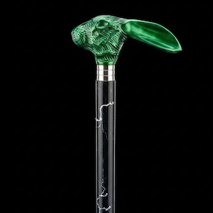 Può includere: Un bastone da passeggio con un'impugnatura a forma di testa di coniglio verde metallizzato con lunghe orecchie. L'impugnatura è fissata a un fusto nero con venature bianche simili al marmo, separato da un collare in metallo argentato.