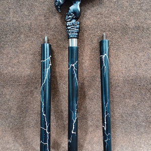 May include: A three-piece walking stick featuring a black handle carved into the shape of a raven perched on a skull. The shaft is black with a white lightning bolt pattern. Each segment includes a threaded metal connector for assembly.