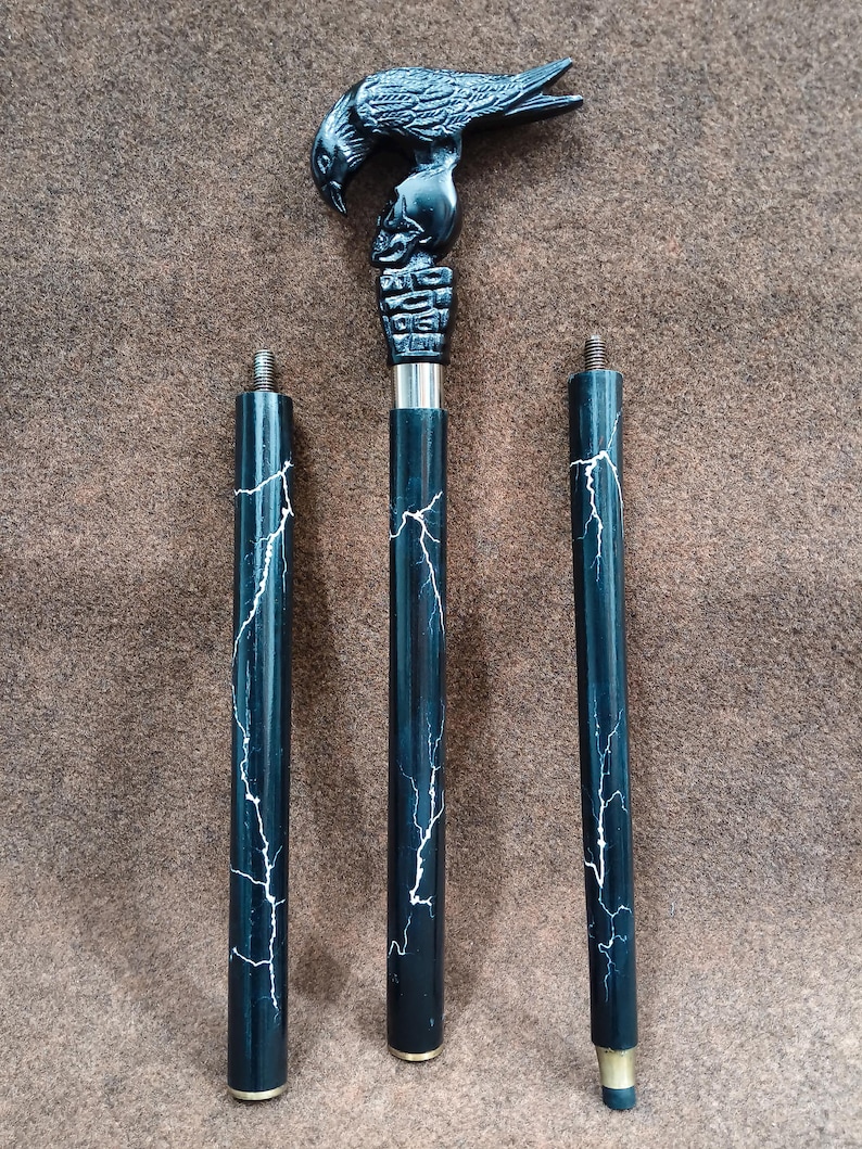 May include: A three-piece walking stick featuring a black handle carved into the shape of a raven perched on a skull. The shaft is black with a white lightning bolt pattern. Each segment includes a threaded metal connector for assembly.