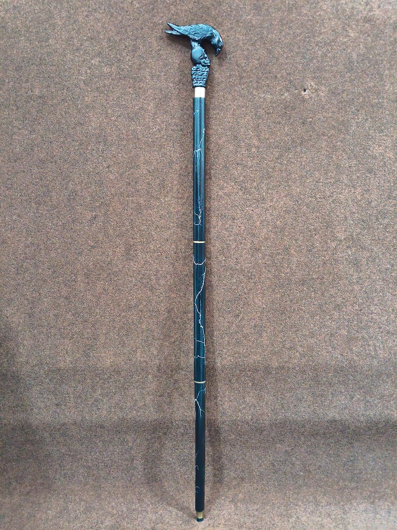 May include: A walking stick featuring a black handle shaped like a bird perched on a skull. The shaft is dark green with thin white marble-like veining and two gold-colored metal bands separating the segments. The base has a small metal tip.