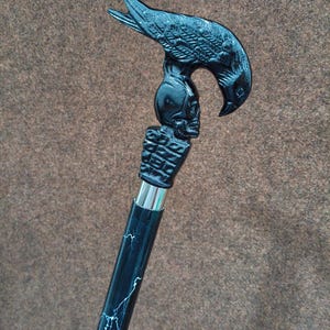 May include: A black walking cane featuring a handle shaped like a bird perched atop a human skull. The handle is connected to a black shaft with white lightning-like streaks by a silver-colored metal collar. The design is monochromatic and detailed.