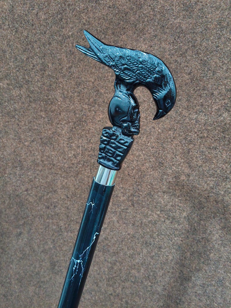 May include: A black walking cane featuring a handle shaped like a bird perched atop a human skull. The handle is connected to a black shaft with white lightning-like streaks by a silver-colored metal collar. The design is monochromatic and detailed.