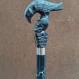 May include: A walking stick featuring a black handle shaped like a raven perched atop a human skull. The handle sits on a textured base above a silver-colored metal collar. The shaft is black with a white lightning bolt pattern running vertically.