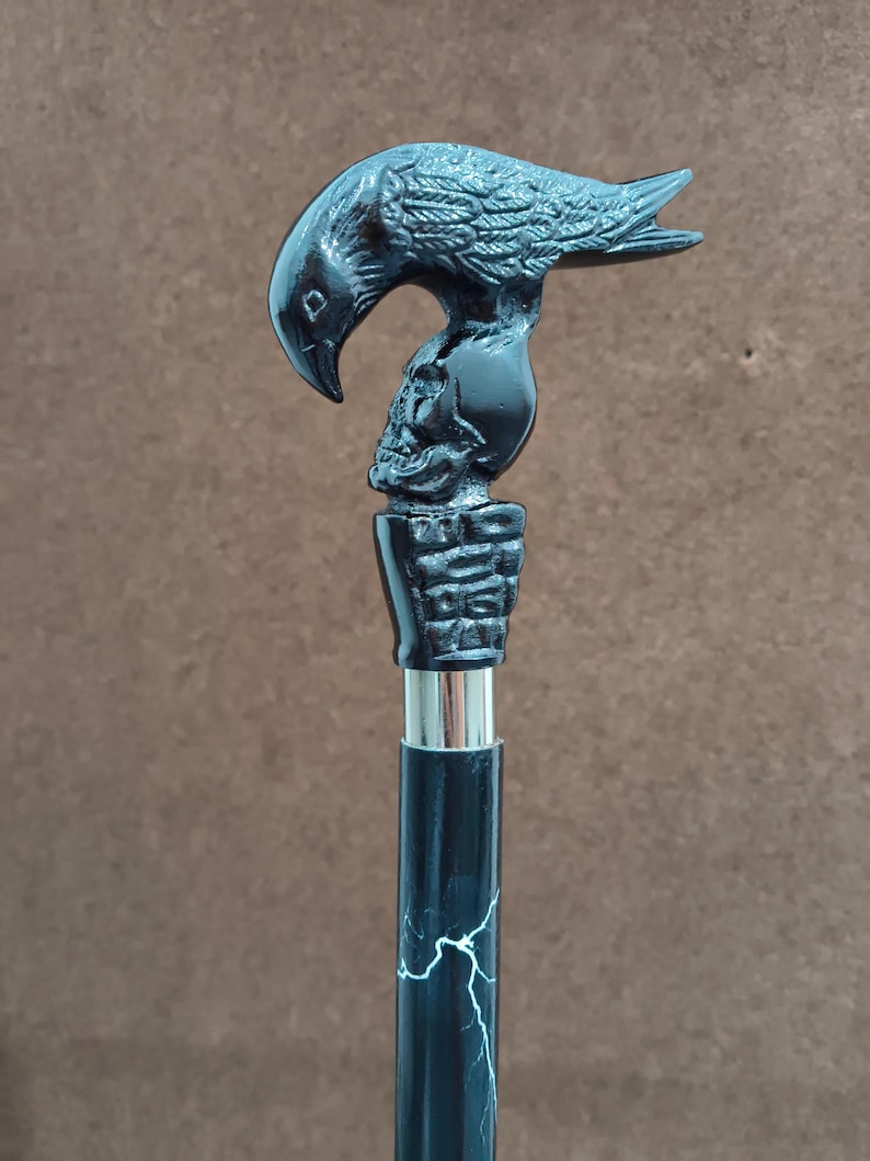 May include: A walking stick featuring a black handle shaped like a raven perched atop a human skull. The handle sits on a textured base above a silver-colored metal collar. The shaft is black with a white lightning bolt pattern running vertically.