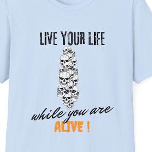 May include: A light blue short-sleeve t-shirt featuring a central graphic of a vertical stack of skulls. Above the skulls is the text "LIVE YOUR LIFE" in black, and below is the text "while you are ALIVE !" with the word ALIVE in orange.