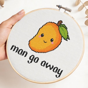 May include: A cross-stitch embroidery project in a round wooden hoop featuring a pixel-art style orange mango with a green leaf and a smiling face, positioned above the black text that reads "man go away" on a white fabric background.