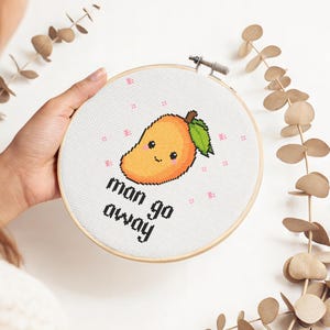 May include: A circular wooden embroidery hoop containing white fabric with a cross-stitch design of a smiling orange mango with a green leaf. The text "man go away" is stitched in black block letters below the mango, surrounded by small pink accents.