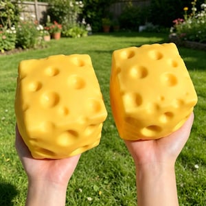 May include: Two yellow, cube-shaped toys resembling Swiss cheese, each with numerous round holes. The toys are held in front of a green grassy background. The cheese-like toys are a bright, cheerful yellow.