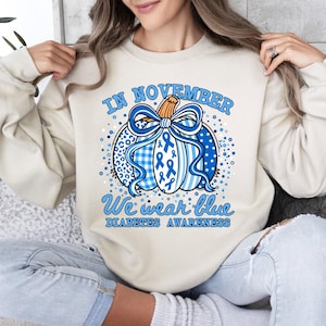 May include: A beige crewneck sweatshirt featuring a graphic of a pumpkin decorated with blue ribbons, leopard print, and plaid patterns. The text reads IN NOVEMBER WE WEAR BLUE DIABETES AWARENESS, surrounded by blue dots and a large blue bow.