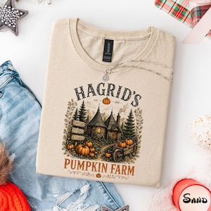 May include: A sand-colored t-shirt featuring a graphic print of a stone cottage, pine trees, and pumpkins. The text reads HAGRID'S est. 1981 PUMPKIN FARM, with smaller signs labeled PUMPKINS, POTIONS, and PLANTS. The design includes orange and green tones.