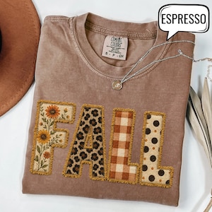 May include: A brown short-sleeved t-shirt with the word FALL appliqued in four distinct patterns: sunflowers, leopard print, brown and white gingham, and brown polka dots. The shirt features a Comfort Colors label and a silver necklace.