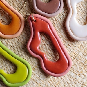 May include: Five pairs of dangle earrings featuring irregular, organic, open-centre shapes. Each earring has a smooth, glossy finish with a two-tone colour scheme, including combinations of lime green, orange, brown, terracotta, and cream with gold-tone hooks.