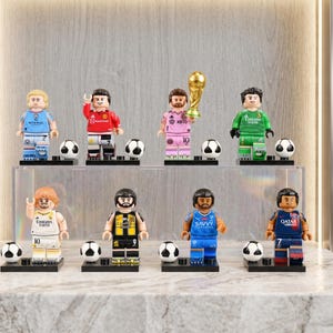 May include: A collection of Lego-style soccer player figurines in various team uniforms. Each figure stands on a black base with a soccer ball. One figure holds a golden trophy. The figurines are displayed on a clear shelf.