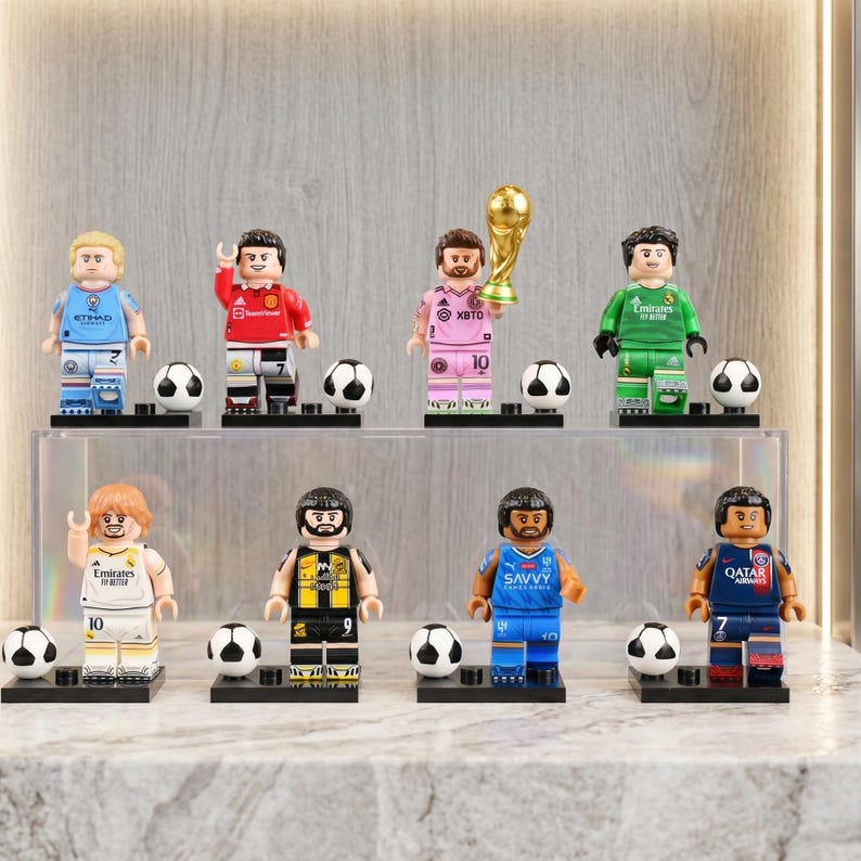 May include: A collection of Lego-style soccer player figurines in various team uniforms. Each figure stands on a black base with a soccer ball. One figure holds a golden trophy. The figurines are displayed on a clear shelf.
