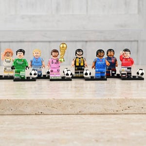 May include: A collection of Lego-style soccer player figurines, each with unique uniforms and accessories. The figurines are standing on black bases, with soccer balls and a golden trophy. The color palette includes green, blue, pink, and red.