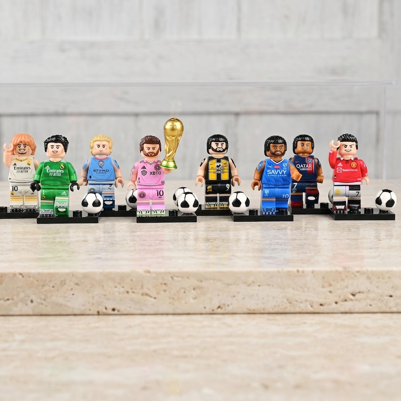 May include: A collection of Lego-style soccer player figurines, each with unique uniforms and accessories. The figurines are standing on black bases, with soccer balls and a golden trophy. The color palette includes green, blue, pink, and red.