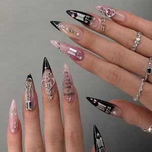 Könnte beinhalten: Ein Satz langer, spitzer Acrylnägel mit verschiedenen Designs. Die Nägel weisen schwarze, rosa und transparente Farbverläufe auf, verziert mit Charms, Strasssteinen und dem BTS-Logo. Einige Nägel haben Text mit den Namen der BTS-Mitglieder.