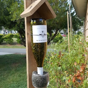 以下が含まれることがあります： ワインボトルに鳥の餌を入れ、ワイングラスを下部につけた木製バードフィーダー。ボトルのラベルには「SOUVERAIN」と「CABERNET SAUVIGNON 2018」と記載されています。