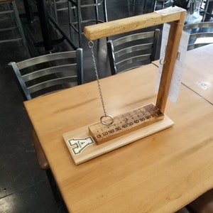 May include: Wooden ring toss game with a numbered scoring track and a metal ring hanging from a chain. The game is set up on a wooden table with a logo that says "A" on the base.