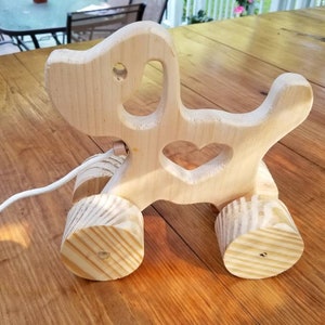 Può includere: Un giocattolo di legno a forma di cane con ritagli a forma di cuore. Il giocattolo ha delle ruote e una corda attaccata.