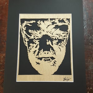 May include: A framed wood cutout of a man's face with a detailed, textured design. The cutout is in a black frame with a white mat. The artist's signature, "Bobby Julin", is in the bottom right corner.