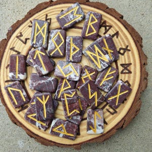 May include: A collection of rune stones with gold symbols, arranged on a wooden slice. The stones are a mix of dark red and white, each displaying a unique symbol. The wooden base has a natural bark edge and some carved symbols.