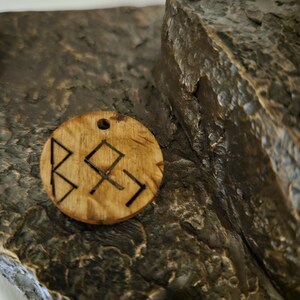 May include: A circular light brown wooden pendant featuring a small hole at the top for suspension. The surface of the wood displays a natural grain pattern and is engraved with three distinct black geometric symbols resembling runic characters.