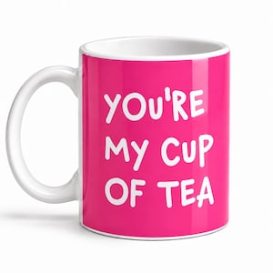 May include: A bright pink ceramic mug with a white handle and interior. The front of the mug features the white text "YOU'RE MY CUP OF TEA" printed in a casual, rounded sans-serif font. The mug has a standard cylindrical shape and a smooth finish.
