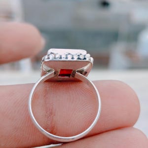 May include: A silver ring featuring a square-cut red gemstone set in a bezel. The metal band has a smooth, polished finish, and the setting is decorated with a series of small, raised silver spheres along the outer edge of the square frame.