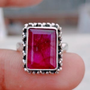 Può includere: Un anello in argento con una grande pietra preziosa rettangolare color rosa intenso con fratture interne visibili. La pietra è incastonata in una cornice d'argento testurizzata e perlata che circonda l'intero perimetro dell'incastonatura.